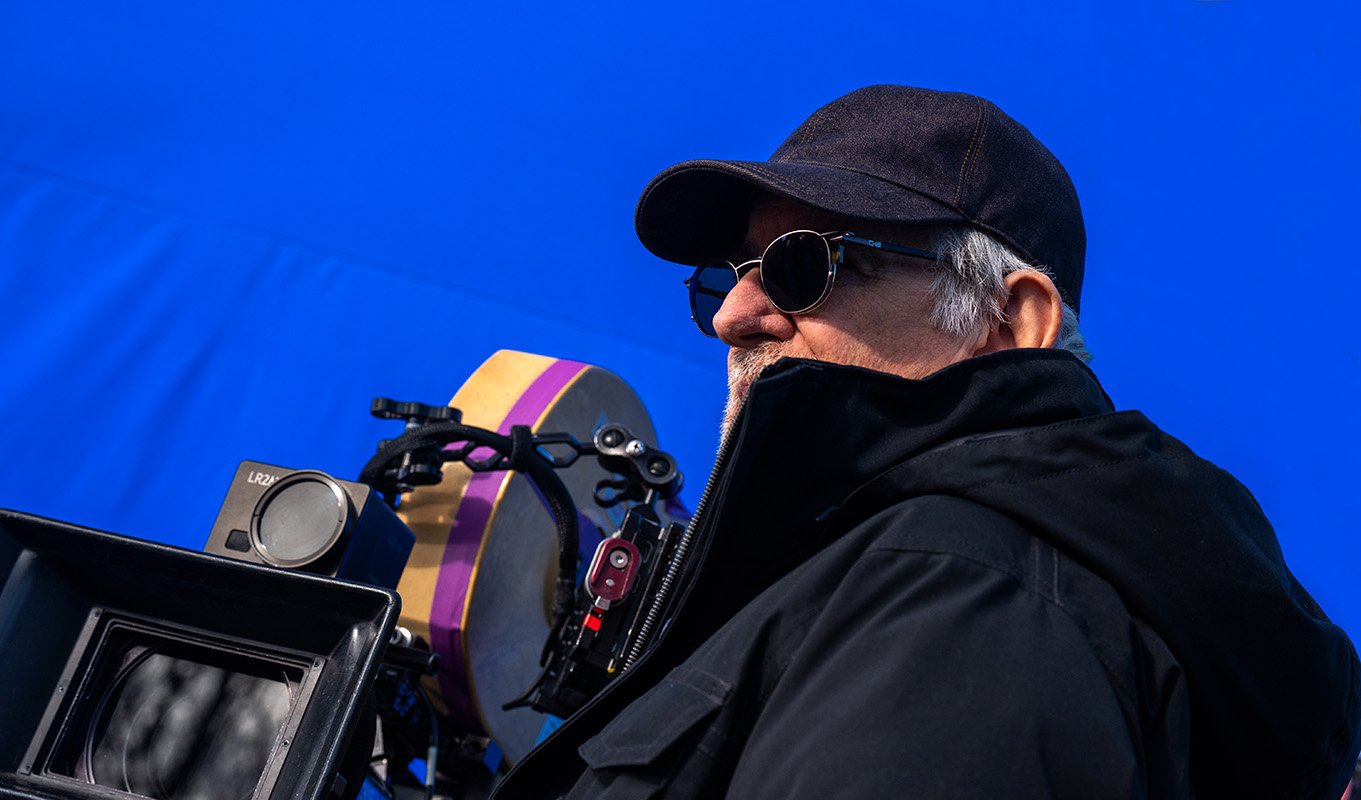 Director Steven Spielberg in a black jacket, baseball cap, and sunglasses looking off-camera next to a professional movie camera on the blue screen set of Disclosure Day.