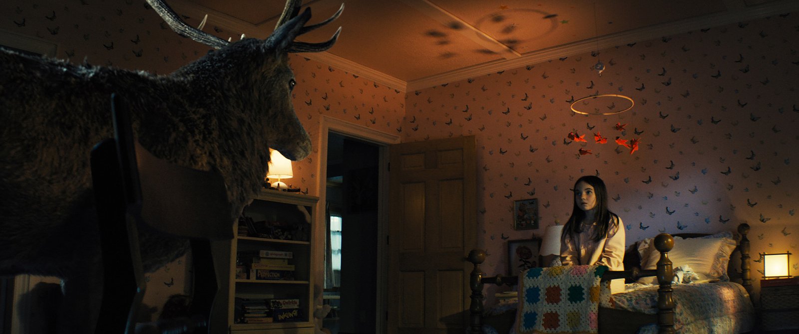 Flashback scene from Steven Spielberg's Disclosure Day 2026 showing child actress young Margaret Fairchild sitting on a bed looking at a giant stag in a dimly lit bedroom.