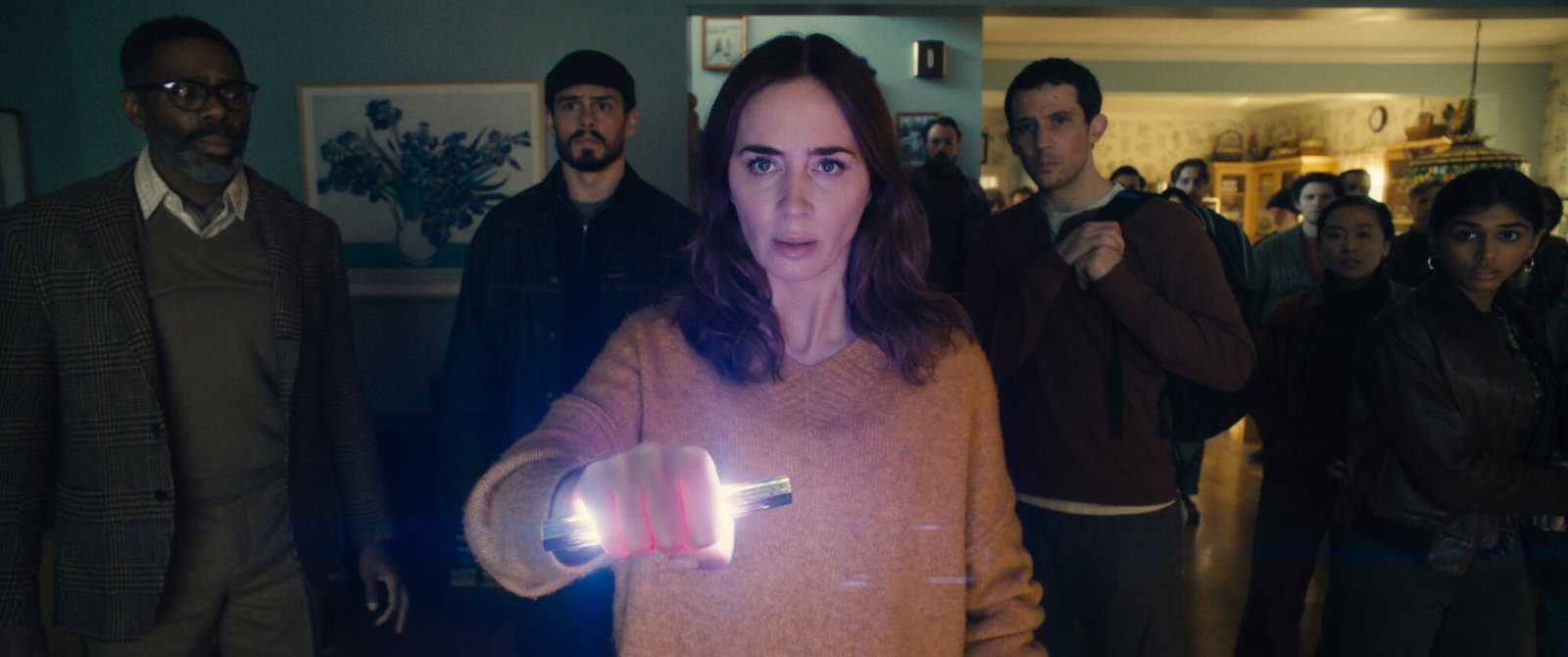 Emily Blunt holding a glowing silver cylinder device forward, surrounded by Colman Domingo, Josh O'Connor, and a crowd of onlookers in a dimly lit room from the movie Disclosure Day.