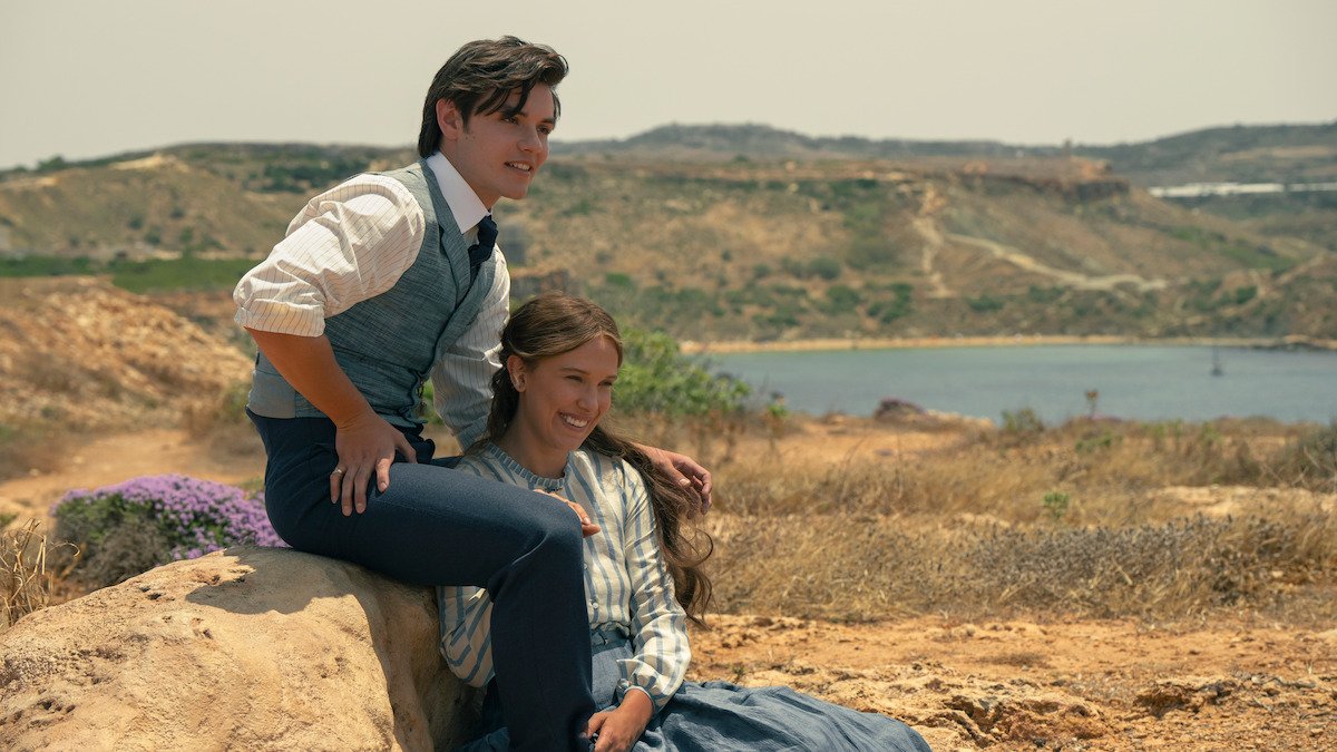 Millie Bobby Brown as Enola Holmes and Louis Partridge as Lord Tewkesbury sitting on rocks by the sea in Enola Holmes 3