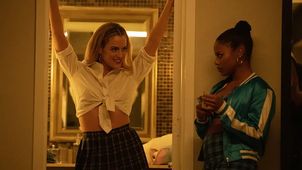 Riley Keough as Stefani with arms raised and Taylour Paige as Zola looking on warily in a motel room in Zola