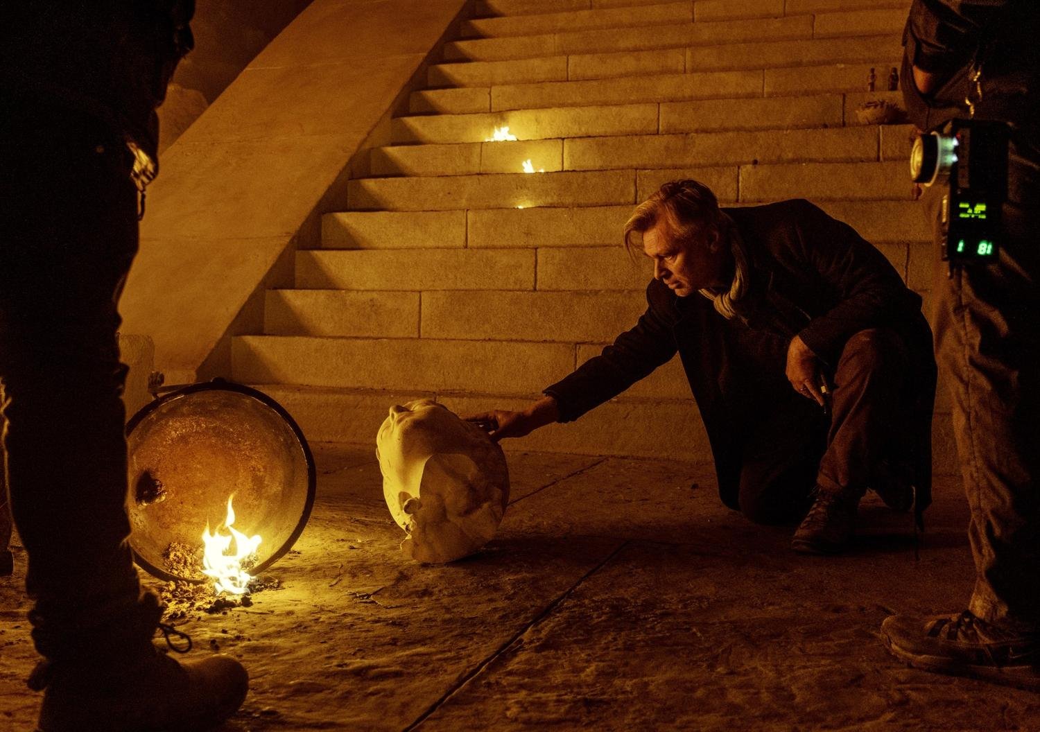 Behind-the-scenes photo from The Odyssey showing the director examining a damaged statue head beside practical fire effects